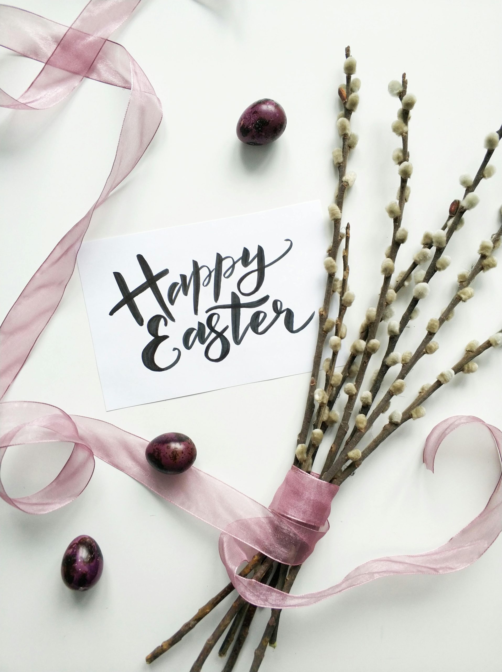 Flat lay of an Easter card with pussy willow branches and pink ribbon on a white background.