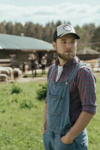 A bearded man wearing a cap and overalls standing in a farmyard on a sunny day.
